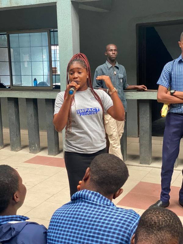 A second fellow in an Impact Project shirt addresses the same group of students with a microphone